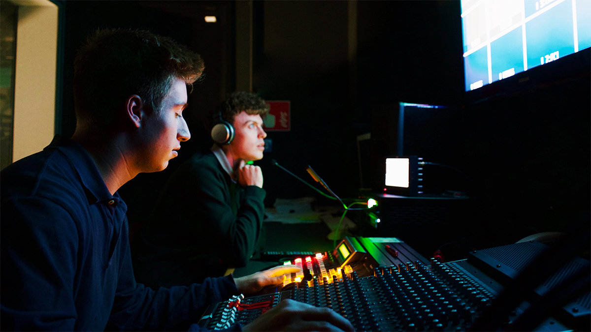 Two students sitting in a dark room with a mixing board and screens