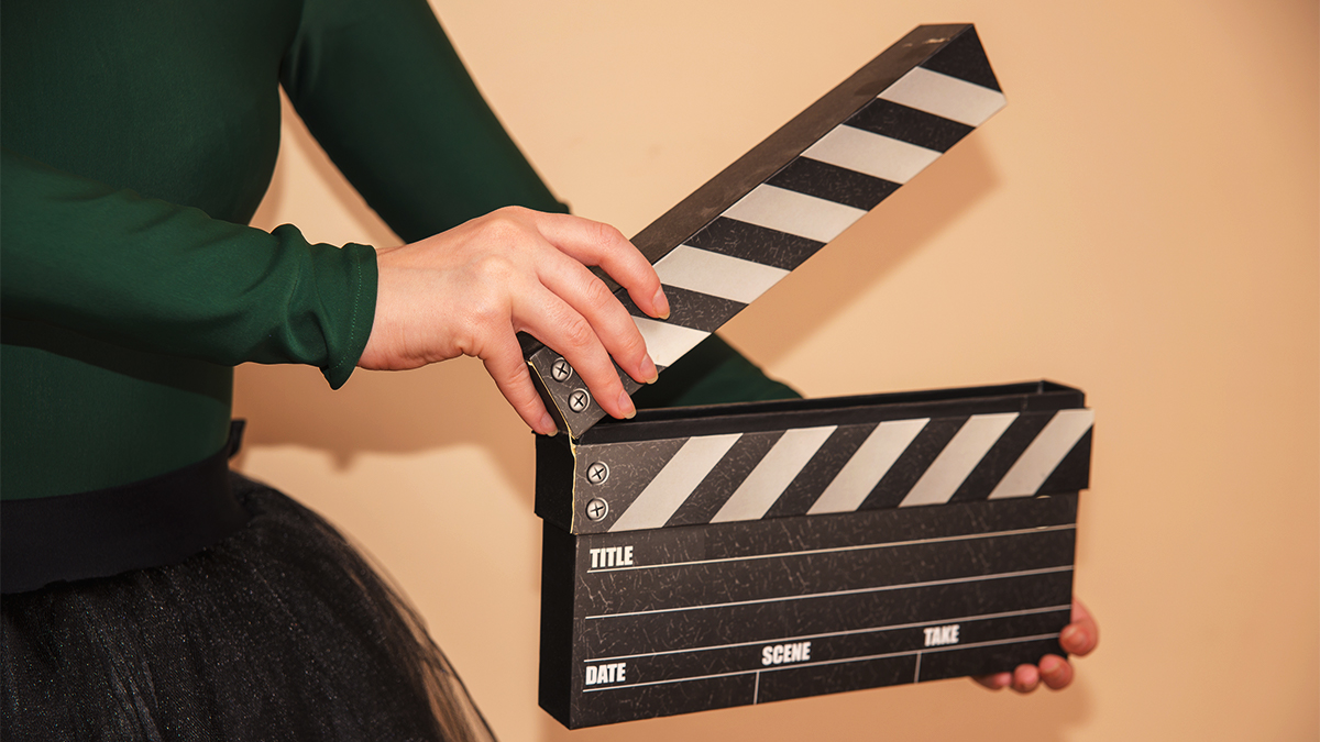 Student in a green top and black skirt holding a film clapperboard, about to close it.