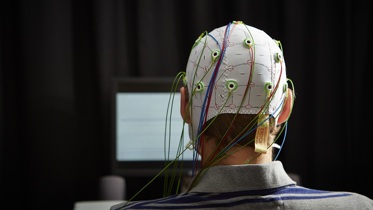 Back of someone's head, they are wearing a cap with wires coming out connected to a computer