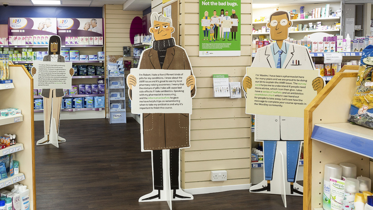Life-size cut outs of illustrated characters holding information on drug-resistant infection in a pharmacy setting