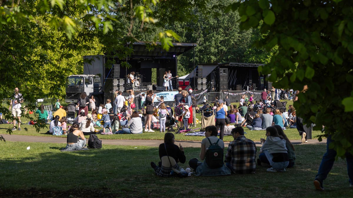 University of Reading, Community Festival 2025. Looking through trees at the stage which is surrounded by a large crowd, sitting in the sun.