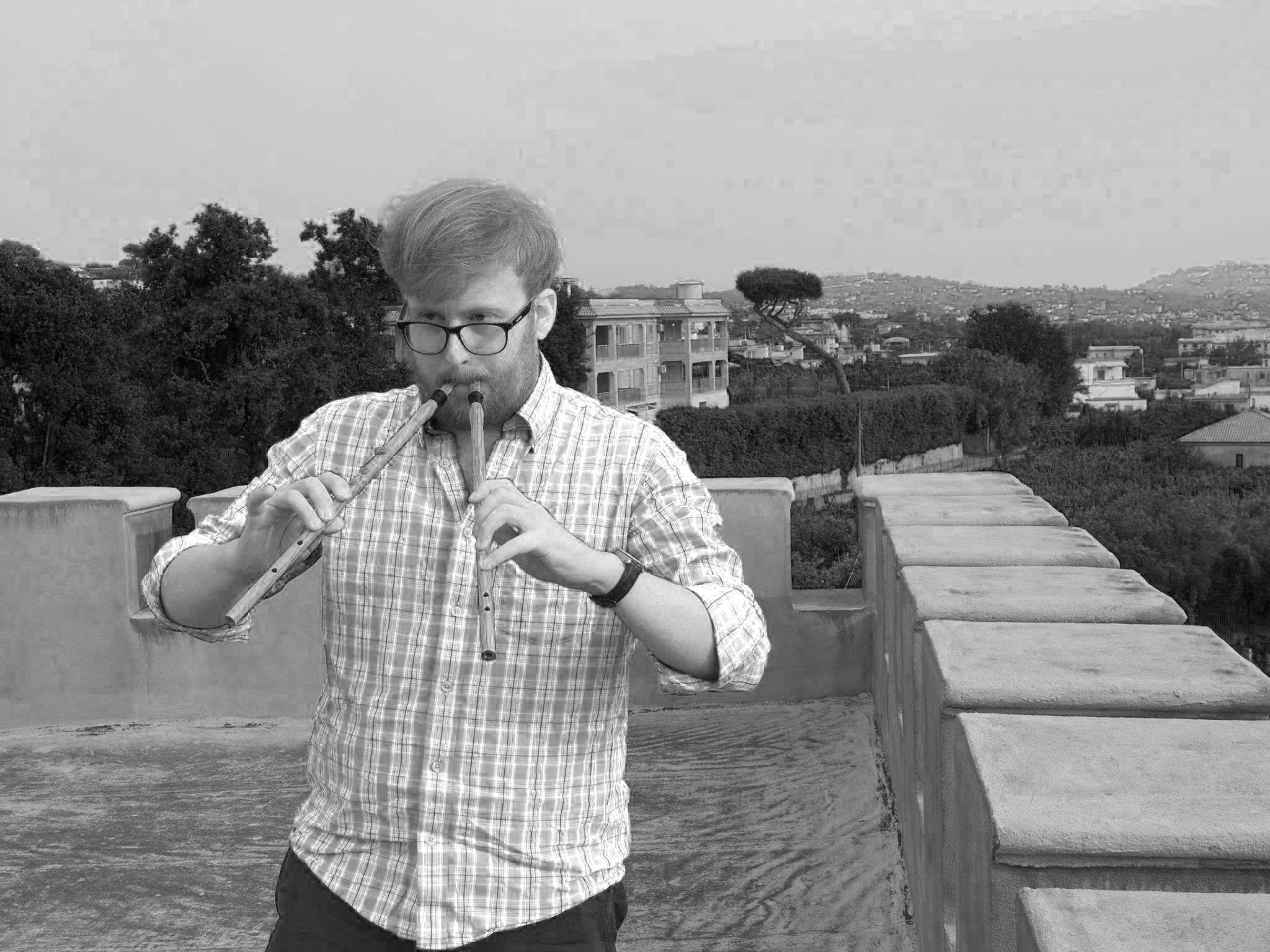 A black and white image  of  a man wearing a checked shirt and glasses, standing on a rooftop playing two flutes
