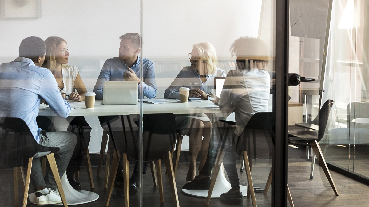 Five people at a meeting sat around a table in a modern office with glass walls