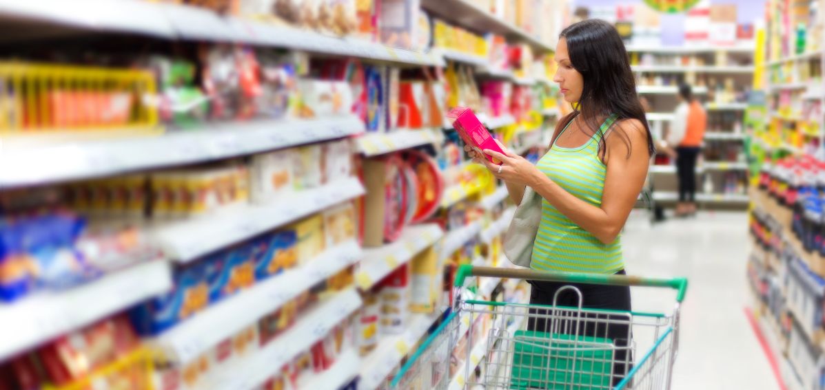 A woman stood with a shopping trolley reading the ingredients on a food packet at the supermarket.