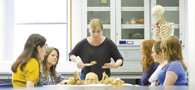 Lecturer and students assembled around a table looking at a human bones. A skeleton stands in the corner.