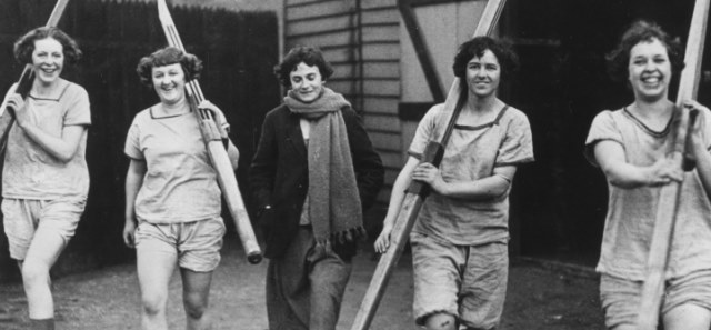 Early to mid 20th century black and white photograph of four female rowers and their cox, carrying their oars.