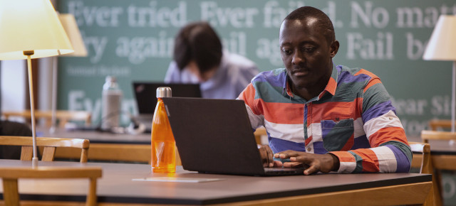 A student sits at a table in the library on his laptop. He is wearing a green, blue, red and white striped shirt. He has an orange water bottle to the side of his laptop. 