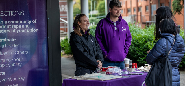 Two students wearing purple RUSU hoodies stand behind a table talking to another student. 