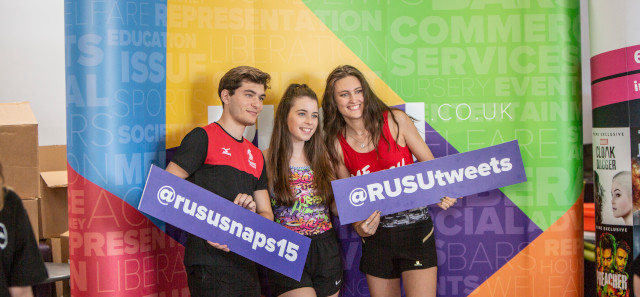 Three students stand in front of a multi-coloured background, holding two pieces of cardboard to promote RUSU social media. One says "@rususnaps15", and the other "@RUSU tweets".