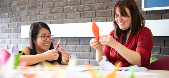Two girls sit smiling whilst doing origami with brightly coloured paper. 