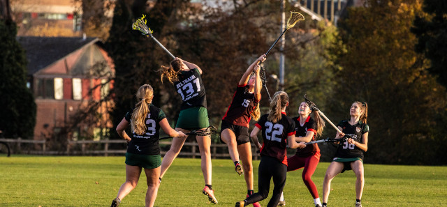 A group of girls are playing lacrosse on the grass. Several are jumping up at the same time with their lacrosse sticks in the air to try and get the ball. 
