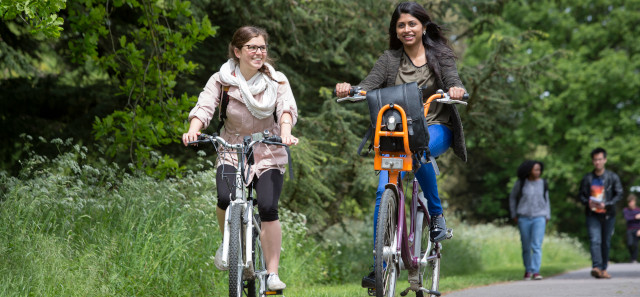 Two students riding bikes against some tall grass. 