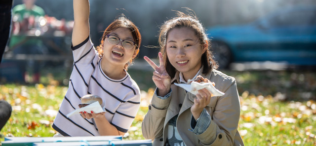Two female students. One has her arm raised and is smiling. The other is doing a peace sign and smiling whilst holding some food. 
