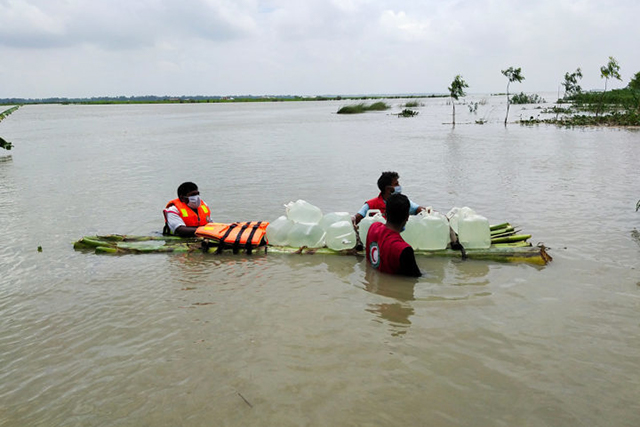 Three men wade through a flooded landscape pulling a raft of clean water bottles