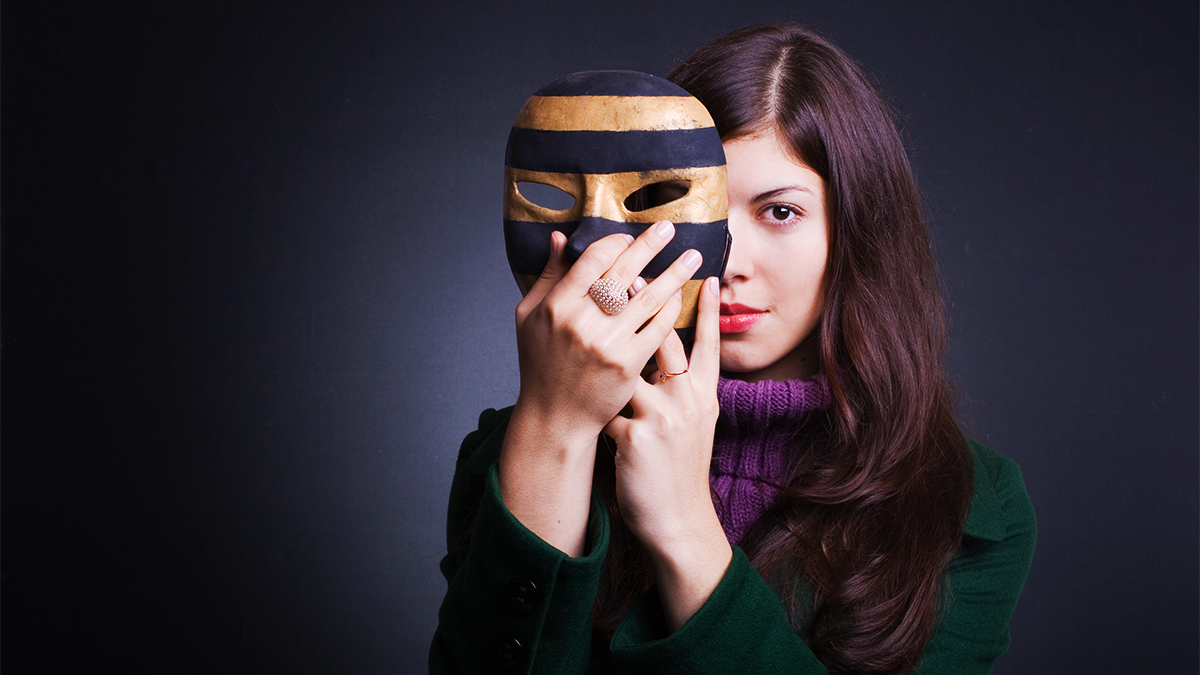 A female student actor with brown hair holding a theatrical mask up partially in front of her face