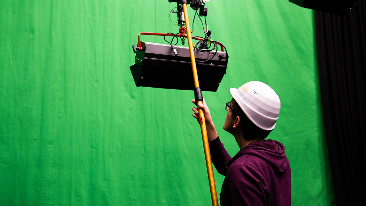 Film student adjusting lighting rig in front of a green screen in Minghella Studios