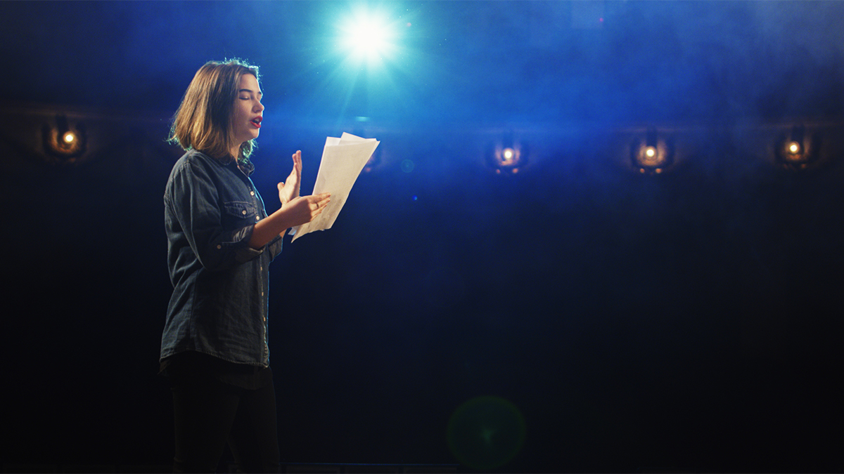 A student on stage facing the audience reading from a script