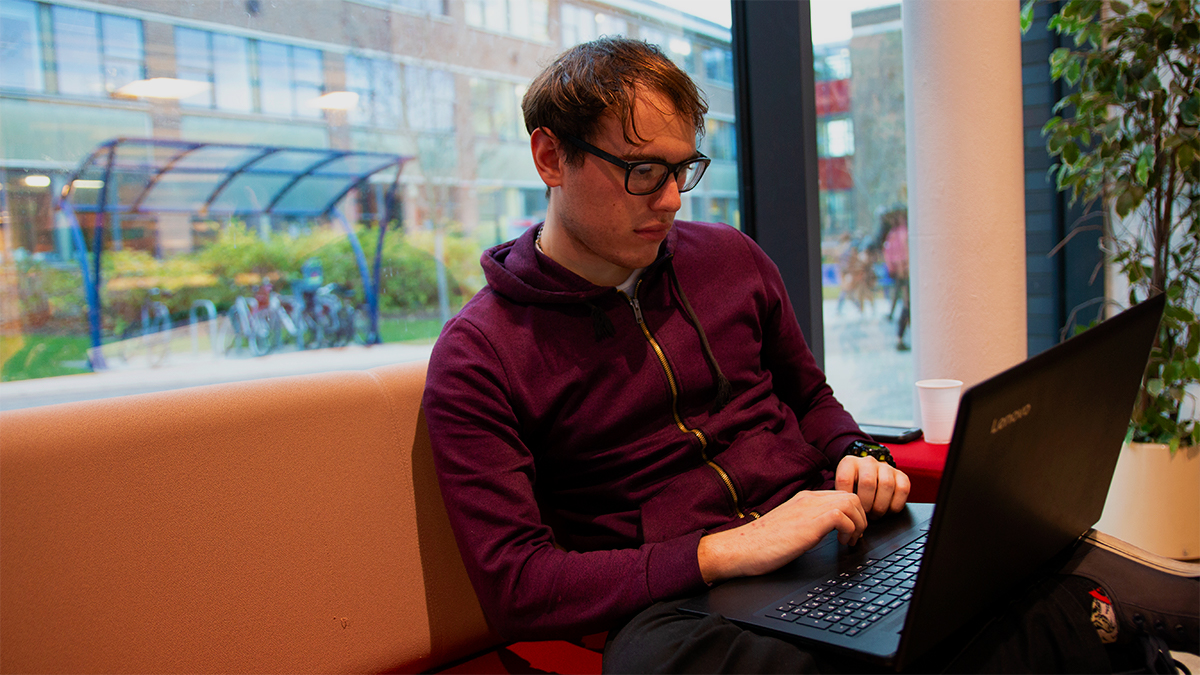 Student sitting on a sofa in Minghella Studios with a laptop with Edith Morley building in the background