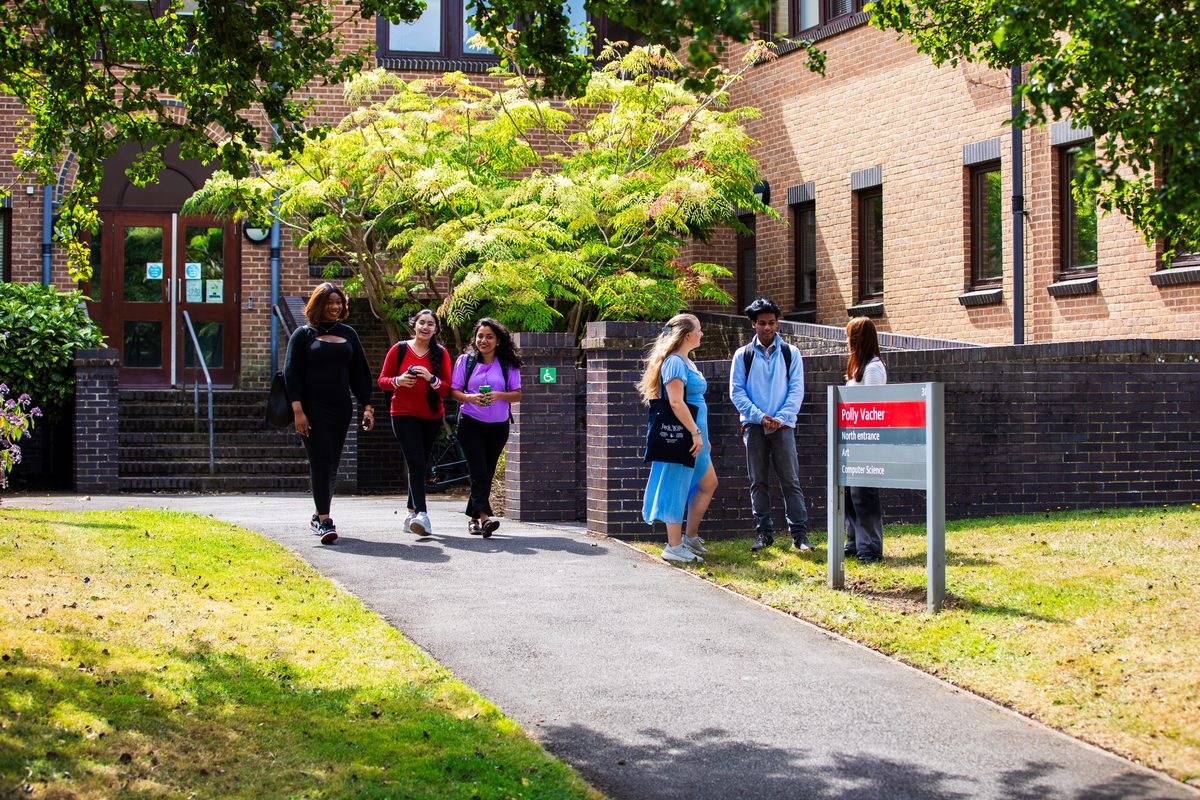 Students walking along a path outside a brick university building surrounded by trees and greenery.
