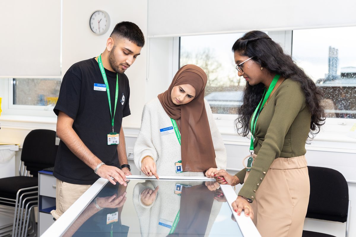Three students wearing ID lanyards using a large touchscreen table together in a bright classroom.