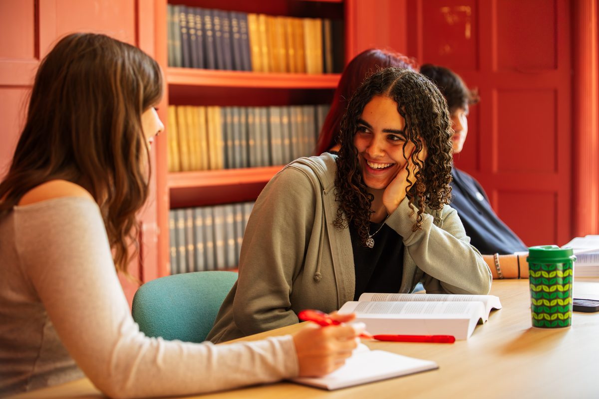 Two students smiling and discussing work while reading books at a table in a library.