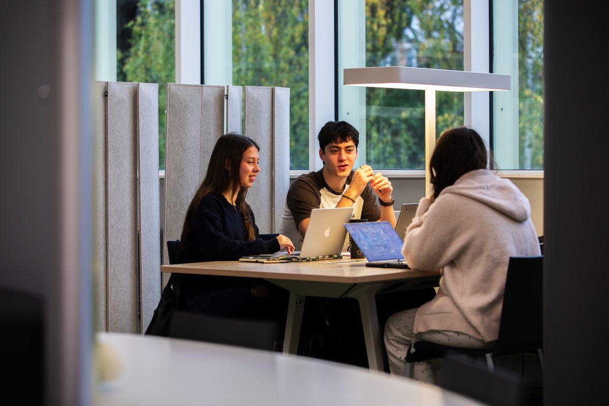 Three students sitting around a table with laptops, talking and collaborating in a study space.