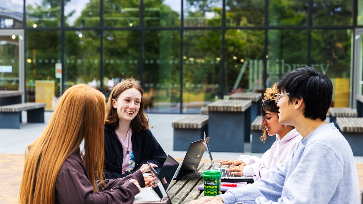 Four students chatting and working with laptops on outdoor table outside the University Library.