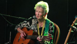 Peggy Seeger, seated against a black background, playing guitar and singing