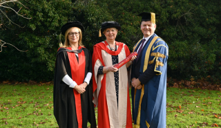 Fiona Talkington in graduation robes holding an honorary degree standing next to Professor Gail Marshall and Vice Chancellor Robert Van De Noort