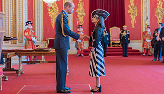 HRH the Prince of Wales presents Deputy Vice-Chancellor Professor Parveen Yaqoob with her OBE. The Prince is in military dress, Professor Yaqoob wears a dark and light striped dress, dark jacket, and hat. They stand in a room with a read carpet and red and gold decoration.