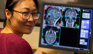 A female technician reviewing images of brain scans. She is turning away from her computer and smiling for the camera.