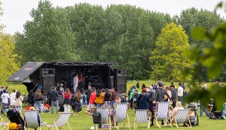 A crowd of people sitting and standing watching a music act on a stage, surrounded by trees and green space