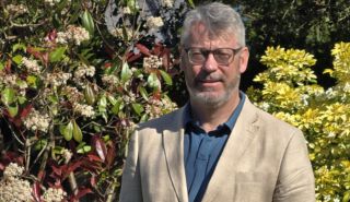 A head and shoulders photograph of Professor Danny Feltham wearing a cream coloured jacket in a garden
