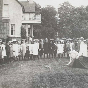 Black and white photo of Eliza Chattaway showing a class of infant children some chicks on the grass. They are wearing Edwardian dress.