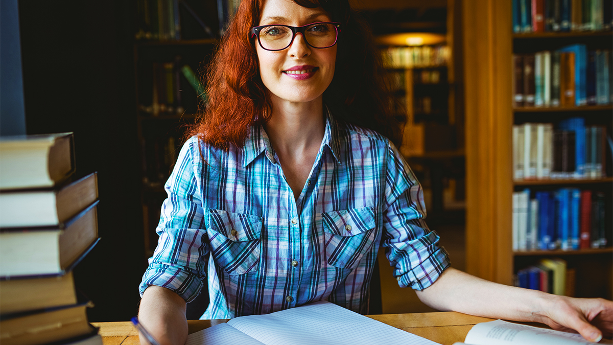 Smiling student sitting in a library with a notebook and a stack of books