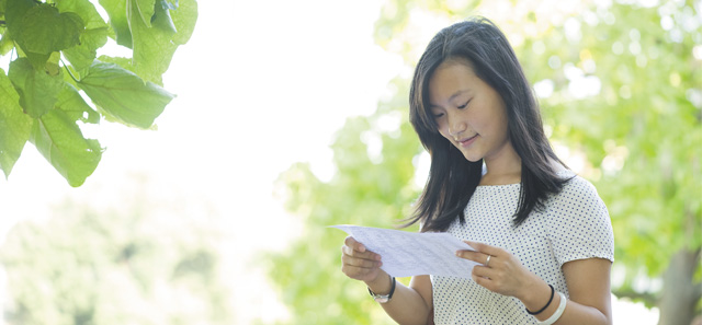 Close-up of a student reading on a sunny day, outside and surrounded by trees.