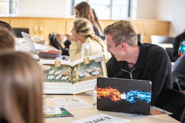 Teachers and students sit at a table with textbooks and laptops