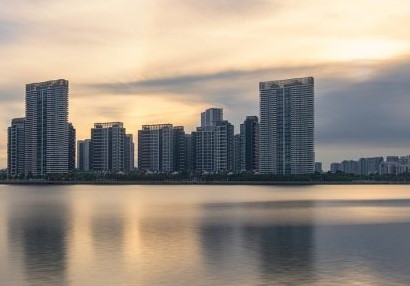 A city skyline under an orange sunset with a calm lake or sea in the foreground