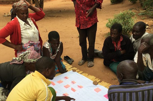 A technical chart or schematic is laid on the ground with eight people gathered around it in discussion